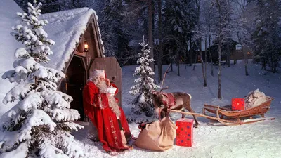 Фото Санта-Клаус / Santa Claus едет в санях, запряженной оленем к  наряженному домику, стоящему среди заснеженных елей, под лунным небом